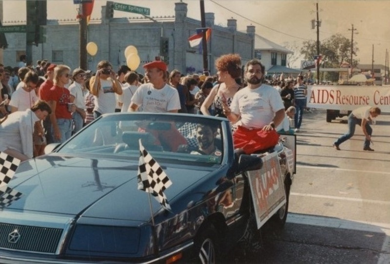 color photograph four people riding in a pride parad float, surrounded by a crows of marchers, banner in the background reads; "AIDS Resource Center"